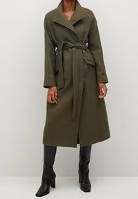 Olijfgroen lange jas met een wikkelontwerp, brede kraag en strikceintuur. Voorzien van twee zijzakken en gemaakt van een textuurstof.
