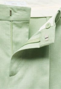 Lichtgroene stof met een gladde textuur; heeft een rits, knoopsluiting en een metalen sluiting; label zichtbaar aan de bovenkant.