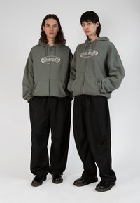 Duos asmenys stovi šalia vienas kito, apsivilkę dideliais, užtraukiamais hoodie alyvuogių spalvos su priekyje esančiu logotipu ir laisvomis juodomis kelnėmis.