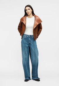 Ženska z dolgimi temnimi lasmi, oblečena v rjavo usnjeno jakno preko bele majice, široke modre kavbojke in črne čevlje, stoji ob svetlem ozadju.