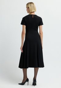 Vestido negro con mangas cortas, falda acampanada hasta la rodilla, detalle de abertura en la espalda, textura lisa. Combinado con tacones altos negros y medias.