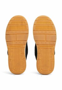 Zapatillas negras con un upper texturizado negro, que presentan una suela de goma amarilla y marrón. "TOMMY JEANS" grabado en la suela en letras mayúsculas.