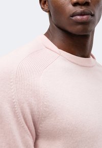 Gros plan d'une personne portant un pull en maille côtelée rose clair, montrant l'épaule, le cou et le bas du visage avec les lèvres fermées.