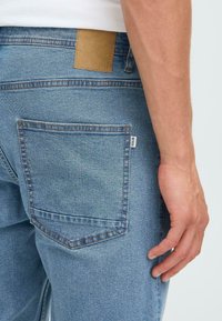 Gai zila džinsa bikses ar aizmugures kabatu ar augsto šuvju un brūnu ādas uzšuvi jostasvietā. Gluda tekstūra ar minimālām nolietojuma pazīmēm.