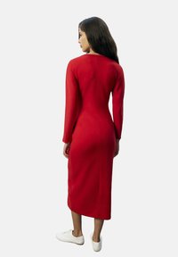 Vrouw met lang, donker haar, gekleed in een aansluitende rode midi-jurk met lange mouwen en witte sneakers, staande tegen een effen achtergrond.
