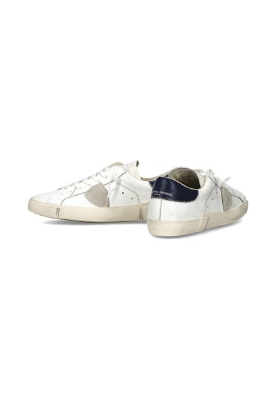 Witte lage sneakers met beige suède accenten, een donkerblauw hielstuk en gebroken-witte zolen, getoond vanuit zij- en achterkant tegen een witte achtergrond.