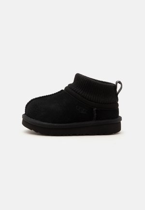Zapatilla negra UGG de tobillo con cuerpo de ante suave, cuello de punto acanalado, lengüeta en el talón y suela gruesa de goma, mostrada de lado.