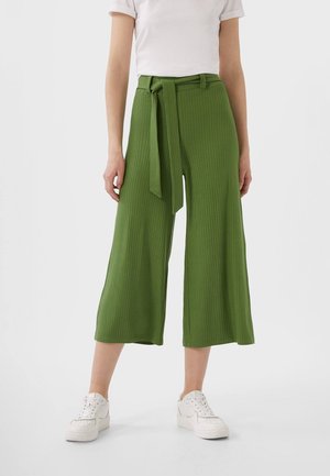 Person iført højtaljede, brede grønne bukser med et matchende stofbælte, hvide sneakers og en hvid kortærmet top mod en ensfarvet baggrund.