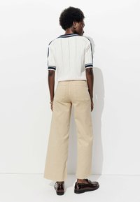 Pull en tricot épais crème avec des rayures bleu marine sur les manches, associé à un pantalon beige à larges jambes, présentant deux poches arrière et un ourlet brut.