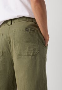 Olive green pants made of cotton, featuring two back pockets with button closures and a small logo detail near the waistband.