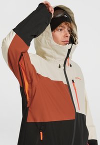 Heren skijas met een color-block ontwerp in beige, bruin en zwart. Gemaakt van waterdicht materiaal met ritssluitingen voor ventilatie en een verstelbare capuchon.