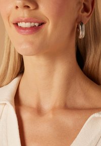 Zilveren creolen met een ribbelstructuur, gedragen door een model met lichte huid en steil blond haar, in een trui met V-hals.