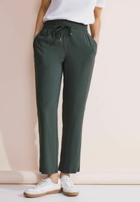 Pantaloni verde, leggeri, con vita elastica e coulisse, dotati di tasche laterali e vestibilità ampia, abbinati a sneaker bianche.
