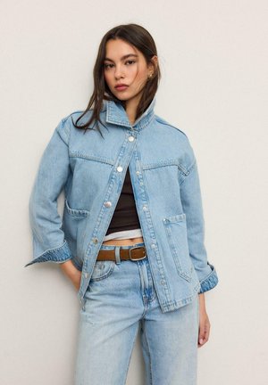 Mlada ženska, oblečena v svetlo modro denim jakno in kavbojke z rjavim pasom, stoji ob enobarvni svetli steni.