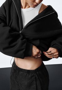 Crna pamučna dukserica s patentnim zatvaračem i ovratnikom podstavljenim runom, nošena preko bijele majice bez rukava, u kombinaciji s tamnim hlačama s elastičnim pojasom. Reflektirajući prstenovi na prstima.