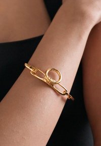 Χρυσό βραχιόλι με κυκλικό βρόχο και σχέδ�ιο κλειδώματος, με ομαλή υφή. Φορεμένο σε ένα χέρι με απόχρωση δέρματος σε αντίθεση με σκούρο φόντο.