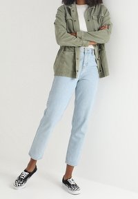 Olive green button-up jacket over a white ribbed top, light blue straight-leg jeans, and black sneakers with white flame pattern accents.