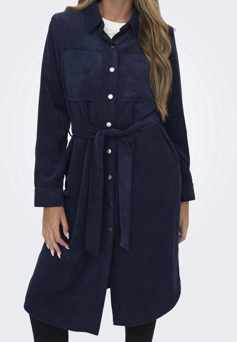 Robe chemise en velours côtelé marine avec un devant boutonné, deux poches poitrine, des manches longues et une ceinture nouée à la taille. Présente une surface texturée.