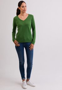 Zelený svetřík s V-výstřihem vyrobený z měkké tkaniny, kombinovaný s přiléhavými modrými džíny a bílými teniskami. Dlouhé rukávy, hladká textura, ležérní design.