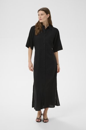 Ženska, oblečena v črno midi obleko z gumbi in kratkimi rokavi, stoji in gleda vstran na enobarvni ozadju.