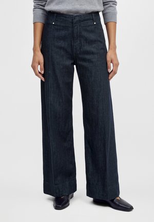 Modèle portant un jean foncé à jambes larges, un pull gris et des chaussures noires sur un fond uni.