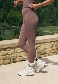 Kõrge vöökohaga lilla retuusid, millel on sile tekstuur ja külgõmbluse detail, kombineeritud valgete spordijalanõude ja mustade triibuliste sokkidega.