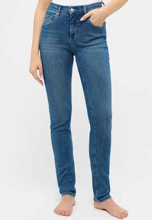 Femme portant un jean skinny taille haute bleu avec bouton et fermeture éclair, pieds nus, debout avec une main dans la poche sur fond blanc.