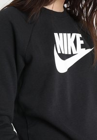 Svart sweatshirt med en stor vit Nike-logotyp över bröstet. Materialet verkar mjukt med ribbad halsringning och en avslappnad passform.