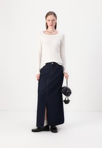 Top bianco a maniche lunghe con coste e scollatura ampia; gonna lunga in denim scuro con spacco frontale e cuciture dorate; scarpe nere in vernice; piccola borsa nera.