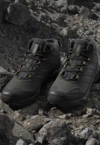 Μαύρα παπούτσια πεζοπορίας Adidas Terrex με χρυσά ρουμπάκια, τοποθετημένα σε πετρώδες έδαφος, που αναδεικνύουν τον ανθεκτικό σχεδιασμό υπαίθριου υποδήματος.