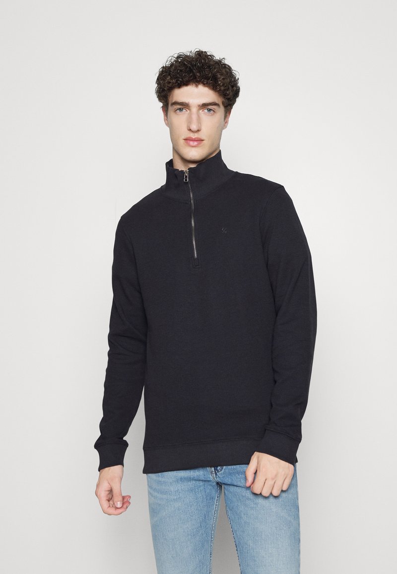 Zwarte rits-hoodie met hoge kraag en gebreide manchetten. Gemaakt van textuurstof, met een klein logo op de borst. Draagt met lichte spijkerbroeken.