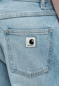 Το ανοιχτό μπλε τζιν παντελόνι διαθέτει πίσω τσέπη με μια λευκή ετικέτα που απεικονίζει ένα μαύρο λογότυπο. Οι ραφές είναι ορατές κατά μήκος των ραφών της τσέπης.