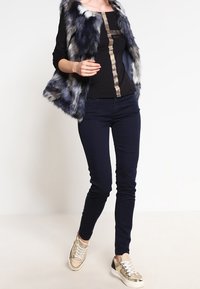 Faux fur-väst i marinblått och grått, med en svart topp som har en metallisk guldkant. Mörkblå slim-fit jeans och sneakers med gulddetaljer.