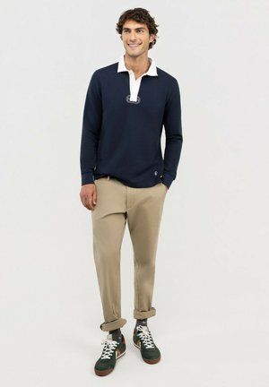 Hombre sonriente con polo azul marino de manga larga, pantalones beige remangados, calcetines grises y zapatillas verdes, de pie con una mano en el bolsillo.