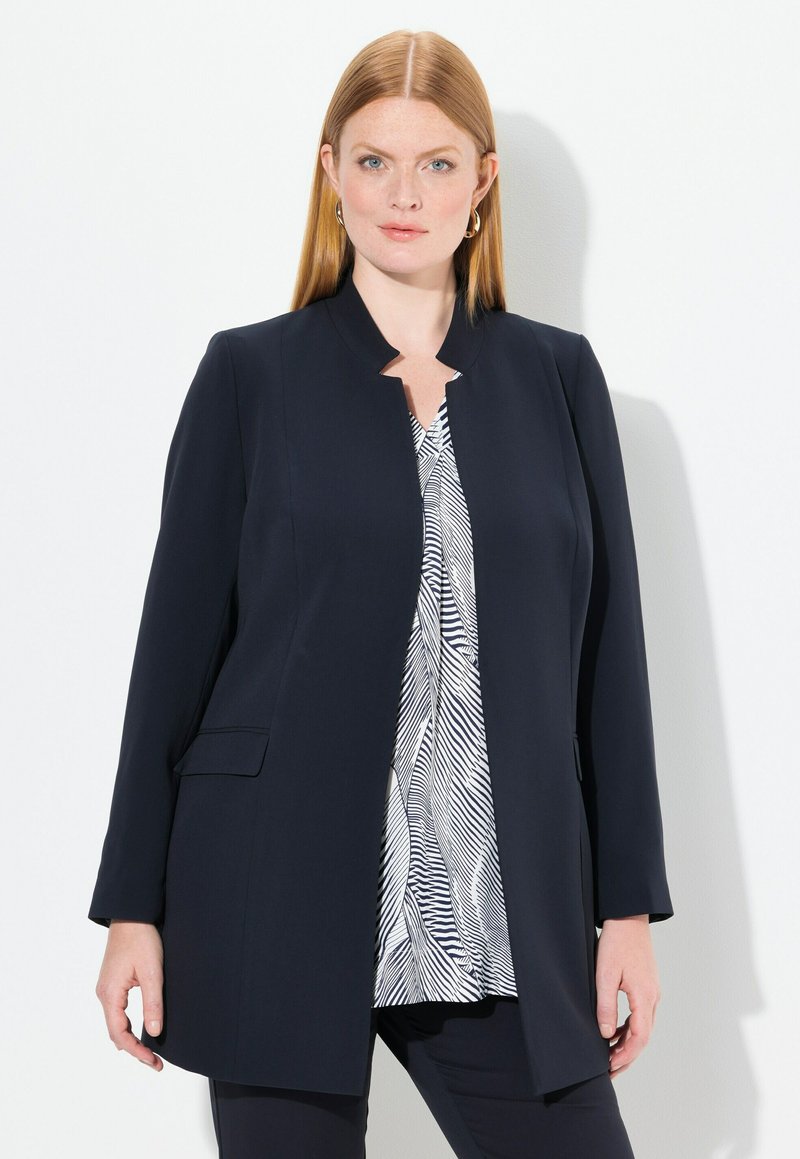 Blazer navy con maniche lunghe, apertura frontale e due tasche laterali. Indossato su una maglietta bianca con un motivo astratto nero. Tessuto di stoffa liscio.