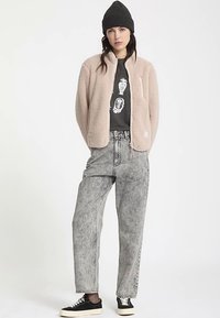 Chaqueta de forro polar en beige suave, cierre de cremallera, usada sobre una camiseta gráfica oscura. Jeans de tiro alto en gris claro y zapatillas negras completan el conjunto.