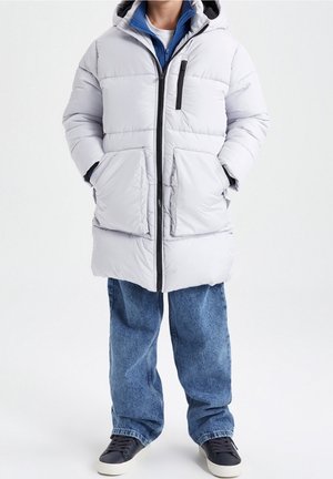 Personne portant un manteau matelassé à capuche gris clair par-dessus une veste zippée bleue, un jean bleu ample et des baskets noires à semelles blanches.