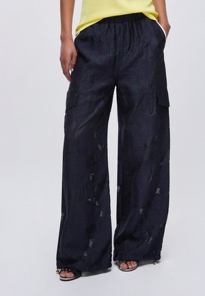 Persona che indossa pantaloni cargo neri a gamba larga con tasche, top giallo e tacchi neri open-toe, in piedi con le mani nelle tasche.