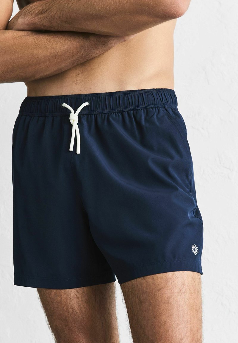 Marineblaue Badeshorts aus leichtem Stoff mit elastischem Bund, weißem Kordelzug und kleinem Logo-Akzent.