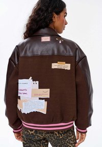 Osoba w brązowej kurtce ze skórzanymi rękawami, różowymi paskami na mankietach, z notatkami i sercowym przypinkiem na plecach, w połączeniu z spodniami w panterkę.