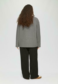 Grauer oversized Pulli mit langen Ärmeln, kombiniert mit schwarzen weiten Hosen und beigen Schuhen, von hinten gezeigt. Lockige Haare sind sichtbar.