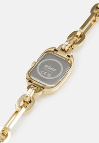 Montre dorée avec un cadran rectangulaire gris, design de bracelet à maillons et dos gravé affichant la marque et les détails de résistance à l'eau.