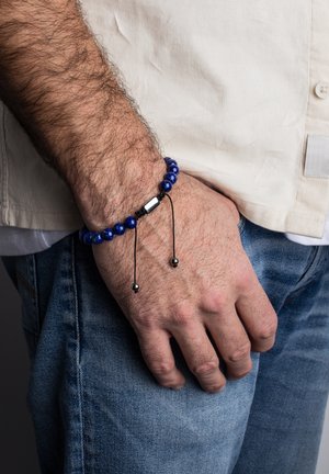 Blue bead bracelet with a silver center detail, adjustable black cord, and two small silver accents, worn on a hand against denim.