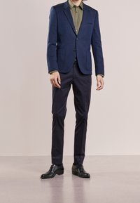 Blazer azul marino con tela texturizada, camisa verde debajo, pantalones oscuros y zapatos negros. El blazer tiene dos bolsillos y un botón frontal.