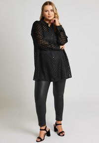 Czarna przezroczysta bluzka w kropki, z długimi rękawami, kołnierzykiem i guzikami z przodu; w połączeniu z czarnymi legginsami z sztucznej skóry i sandałami na obcasie.