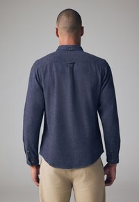 Camisa azul con textura, de manga larga y cuello con botón. Cuenta con botones marrones en los puños y un pliegue en la parte trasera para mayor libertad de movimiento.