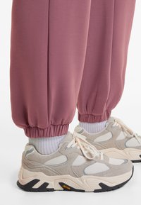 Lyserøde træningsbukser med elastiske manchetter, parret med beige og hvide sneakers, der har mesh- og ruskindsstrukturer samt en sort Vibram-sål.