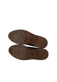 Semelles de chaussures en cuir brun avec une texture à motifs et le logo "CRICKIT" en relief. Forme arrondie avec des clous métalliques le long des bords pour le détail.