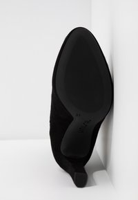 Zapato de tacón alto de ante negro con punta afilada, en posición vertical, mostrando la suela texturizada y la marca de tamaño 37, sobre un fondo blanco.