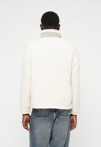 Veste en polaire blanche et duveteuse avec un col haut, surface texturée et longues manches, associée à un jean en denim bleu clair.